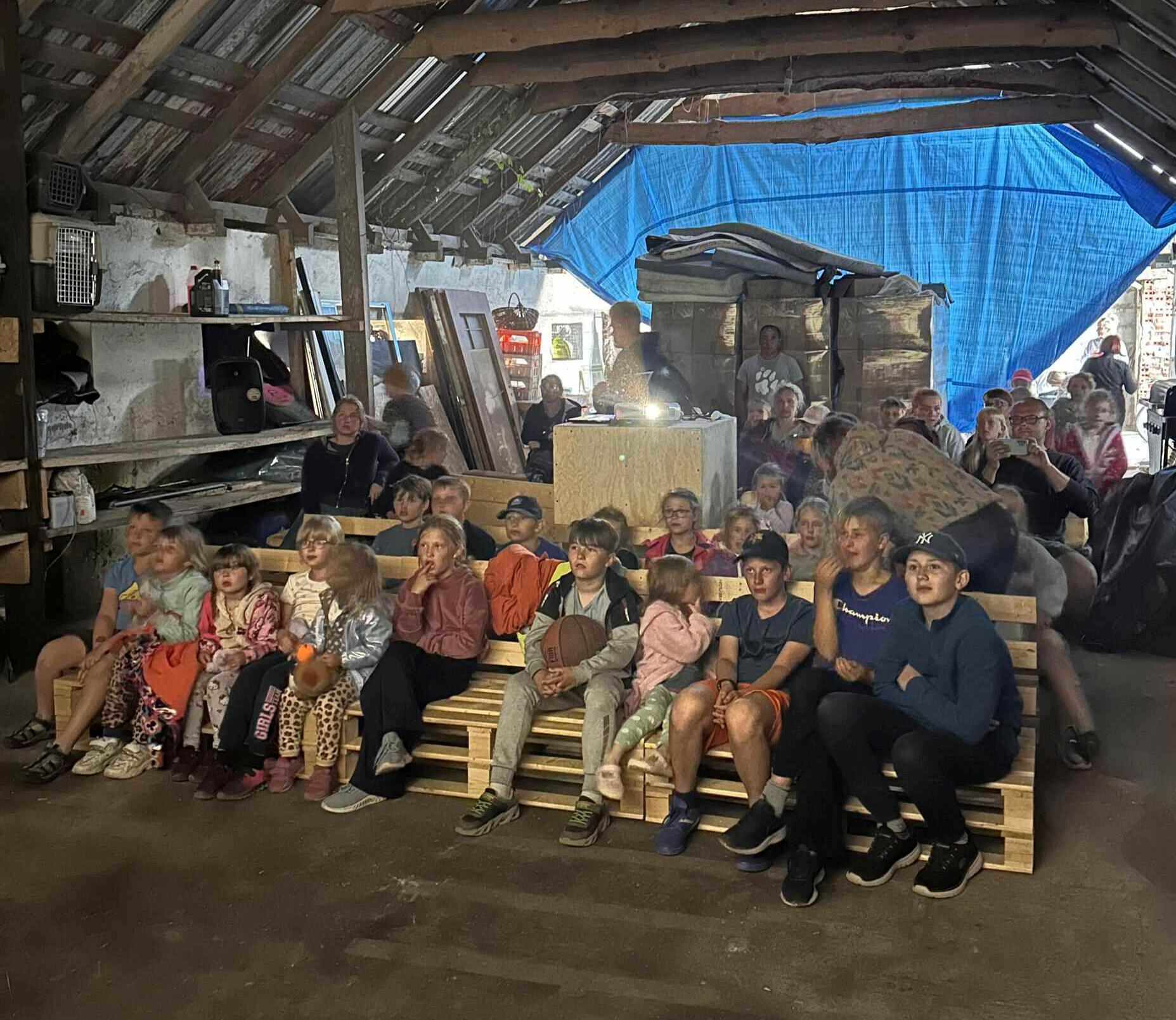 Kino pod dachem stodoły. Grupa dzieci i dorosłych siedzi na paletach w klimatycznej stodole, oglądając film podczas obozowego seansu kinowego.